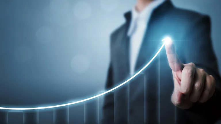 Symbolbild: Mann im Anzug zeichnet mit dem Finger eine steigende Kurve auf einem Balkendiagramm nach.
