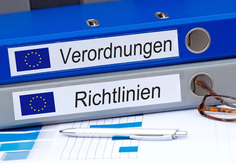 Zwei blaue Aktenordner mit EU-Flaggen und den Aufschriften "Verordnungen" und "Richtlinien" liegen auf einem Schreibtisch. Daneben befinden sich eine Brille, ein Kugelschreiber und ausgedruckte Dokumente mit Diagrammen – Symbolbild für europäische Gesetzgebung und Büroarbeit.