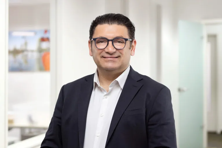 Mann, mittleren Alters, kurze, schwarze Haare, Brille mit rundem Rahmen, tr&auml;gt Sakko und Hemd, l&auml;chelt.