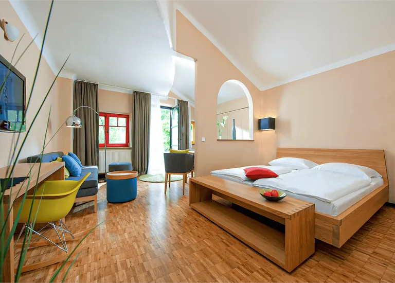 Blick in ein Hotelzimmer. Bett, weiße Bettwäsche, olivgrüne Vorhönge, blaues Sofa